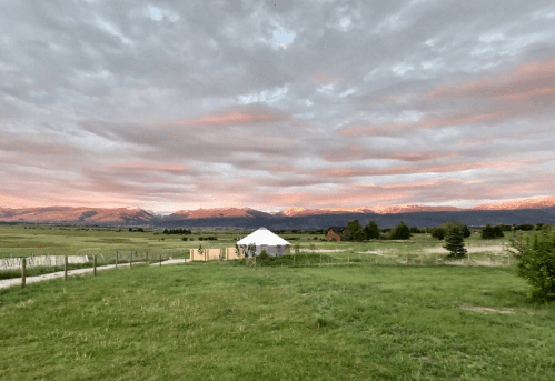 A serene landscape featuring a white tent, green fields, and mountains under a colorful sunset sky.